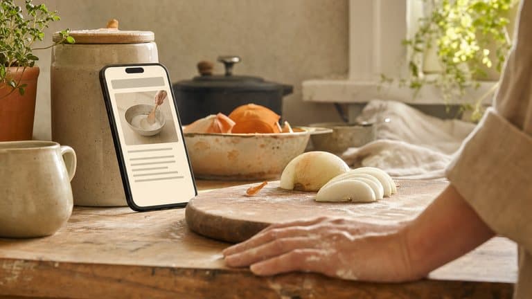A phone propped against a jar on a kitchen counter showing a recipe step, beside a floury board of half-sliced onion